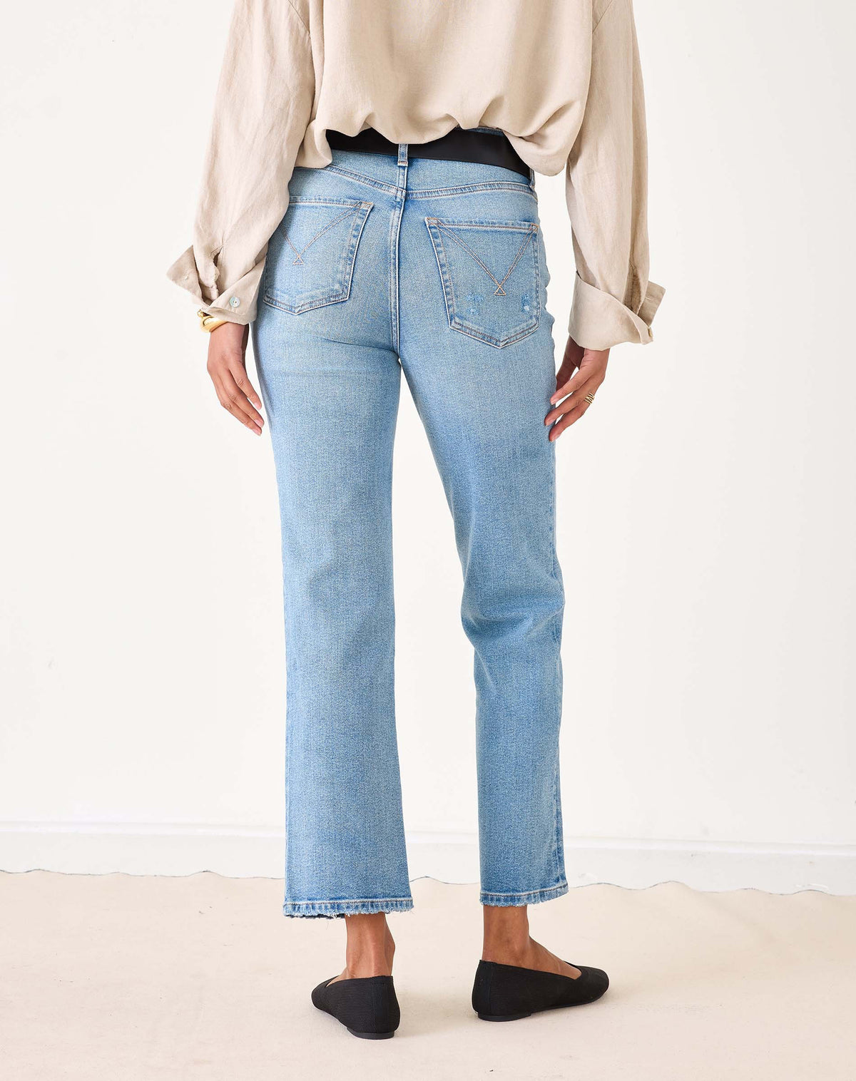 A person wearing a beige button-up shirt, MERSEA Bessie Straight Leg Jean in Caribbean made from recycled cotton, and black flats stands holding a woven straw tote bag with black handles. Their left hand is in their pocket, face not visible.