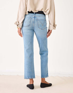 A person wearing a beige button-up shirt, MERSEA Bessie Straight Leg Jean in Caribbean made from recycled cotton, and black flats stands holding a woven straw tote bag with black handles. Their left hand is in their pocket, face not visible.