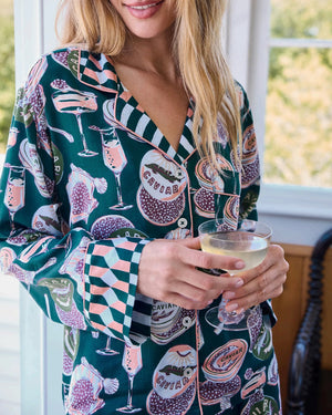 A woman stands barefoot on a wooden floor against a white wall, wearing the PRINTFRESH - Champagne & Caviar Eco Satin Soho Set in Peacock Feather by PRINTFRESH, a matching dark loungewear set adorned with colorful floral and fruit motifs and finished with checkered cuffs.