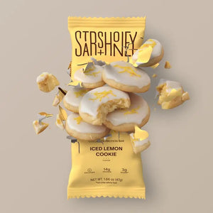 A STARS+HONEY - ICED LEMON COOKIE collagen protein bar in a vibrant yellow wrapper, featuring cookies adorned with white icing and gold sprinkles—some whole, some crumbling—bursting outward against a beige backdrop, capturing a healthy snack from STARS+HONEY packed with delicious flavor.