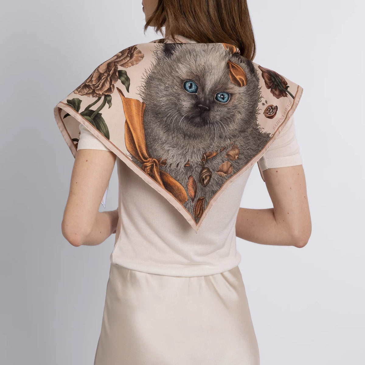 A person in a cream-colored shirt and skirt is seen from the back, wearing a SABINA SAVAGE - RING A RING O' ROSES silk twill triangle scarf by SABINA SAVAGE ENGLAND, featuring a fluffy Ragdoll kitten with blue eyes, framed by English garden flowers.
