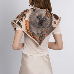 A person in a cream-colored shirt and skirt is seen from the back, wearing a SABINA SAVAGE - RING A RING O' ROSES silk twill triangle scarf by SABINA SAVAGE ENGLAND, featuring a fluffy Ragdoll kitten with blue eyes, framed by English garden flowers.
