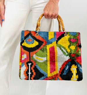 A person in white pants holds the BALDIZ - AVERY BAG IN PALOMA WITH BAMBOO HANDLES, featuring a colorful abstract pattern, against a plain background.
