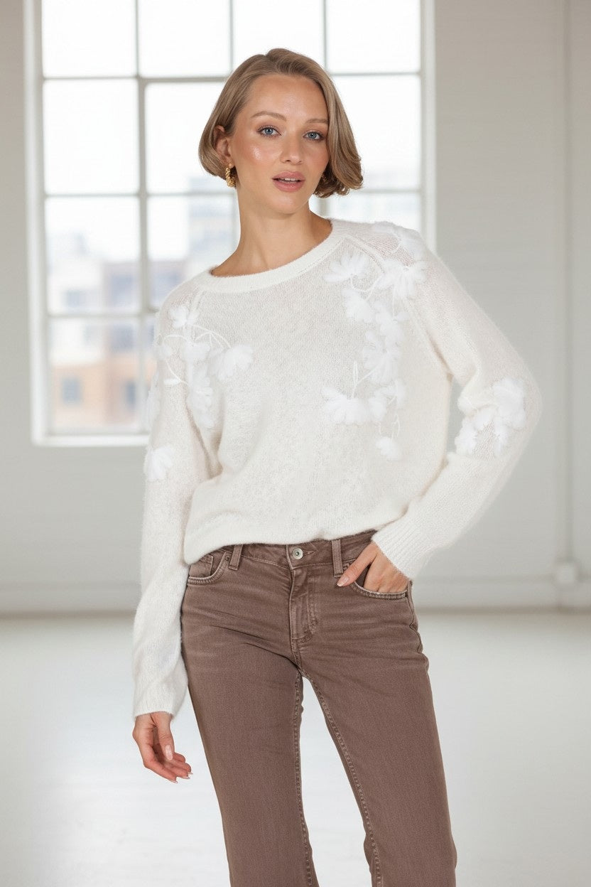 A woman with short blonde hair is seen indoors in front of large windows, looking at the camera with one hand in her pocket. She is wearing brown pants and the JESSIE LIU Merino Wool and Cashmere Blend Pullover with Raglan Sleeve and Applique in Ivory by JESSIE LIU.