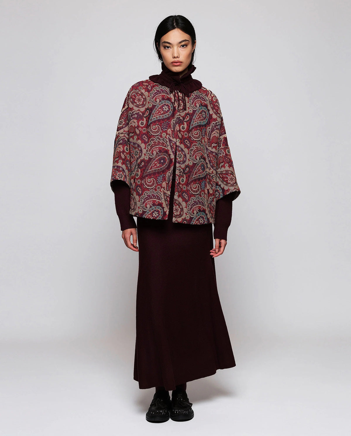 A woman stands against a plain background wearing the MIRTO 1956 Burgundy Paisley Jacquard Jacket—an oversized, cotton-blend piece in rich red and purple tones from MIRTO 1956—styled over a long burgundy dress and black shoes. The relaxed fit of the jacket complements her straight dark hair and neutral expression.