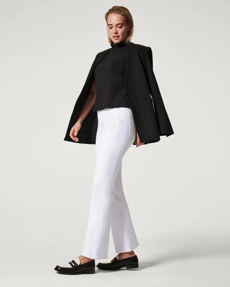 A woman stands in profile against a plain light background, wearing a black blazer draped over her shoulders, a black top, and the SPANX ON THE GO KICK FLARE pant—crafted with Ultimate Opacity Technology for comfort—paired with black loafers.