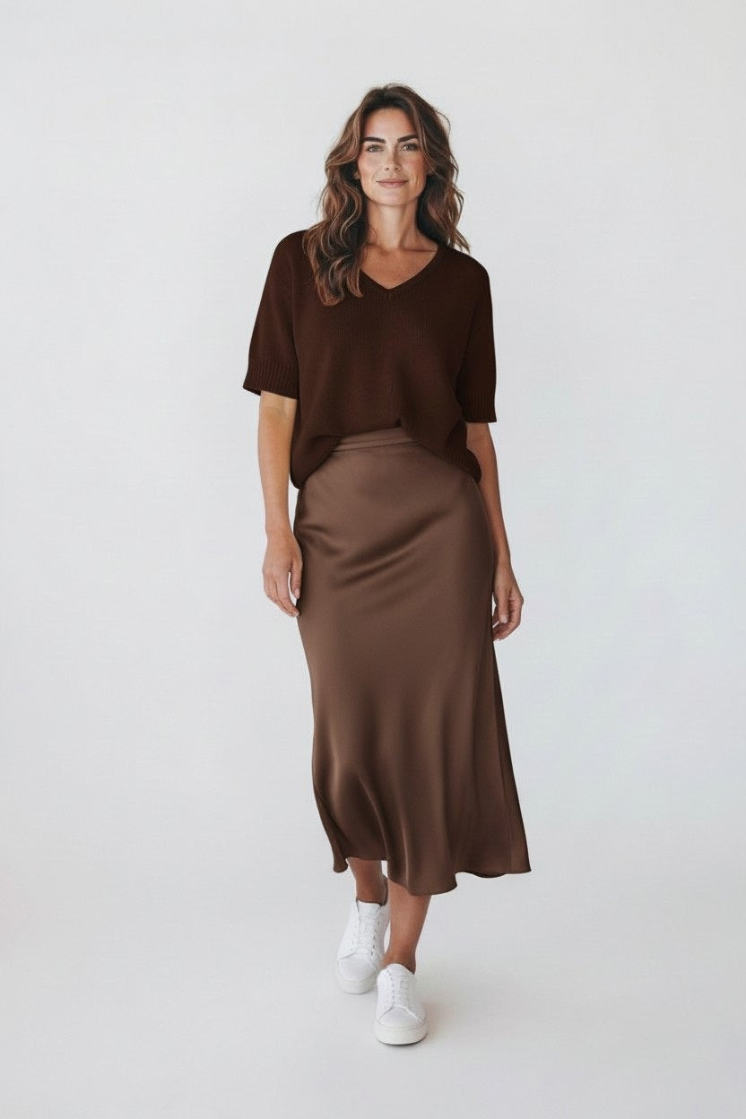 A woman with wavy brown hair smiles against a plain light background, wearing a short-sleeve dark brown top, white sneakers, and the ROSE & CROWN ROSE AND CROWN - SATIN MIDI SKIRT featuring an elastic waistband.