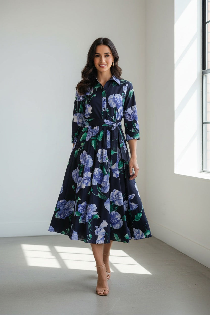 A woman stands with her back to the camera, wearing the elegant LAVI - SPRITZ BLUE POPPIES DRESS by LAVI, featuring a stunning blue midi silhouette decorated with purple and blue poppy flowers. She has short dark hair, gold earrings, and open-toed heels against a plain white background. This striking piece is beautifully made in Italy.