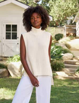 Wearing the FRANK & EILEEN MONTECITO Shell Mock Neck Sweater Shell in Ivory, a person with curly hair and white pants stands outdoors with their back to the camera, in front of a white building surrounded by greenery.