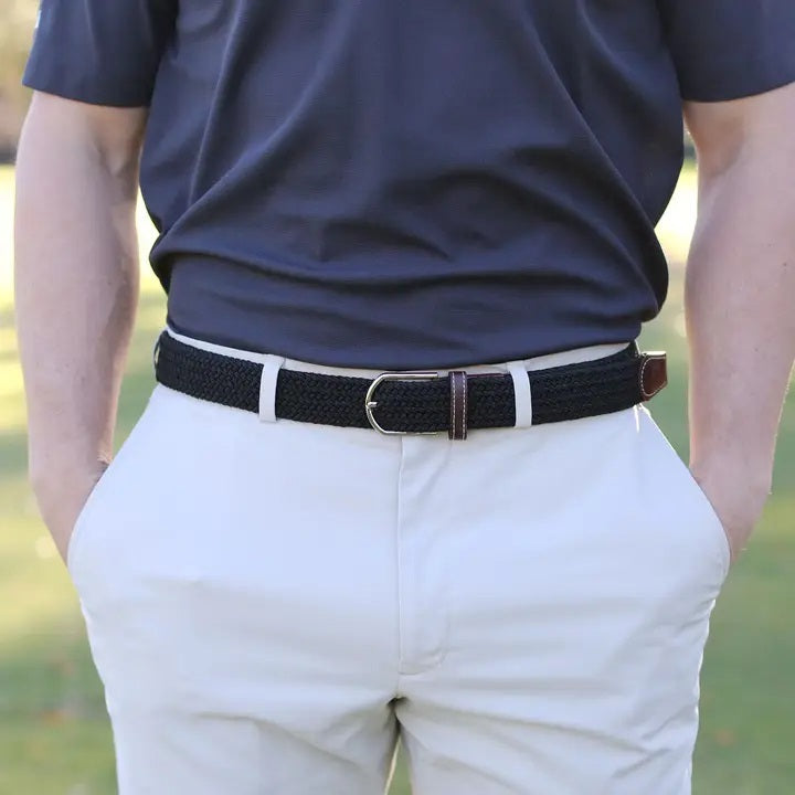 Outdoors, a person with hands in pockets wears a navy blue shirt, light-colored pants, and the ROOSTAS - BETHPAGE WOVEN STRETCH BELT by ROOSTAS featuring brown leather accents.