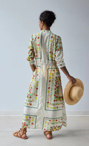 A person stands facing away in the HOPE FOR FLOWERS Band Collar Shirtdress in Custard Floral, paired with brown sandals and holding a straw hat, against a plain light gray background.