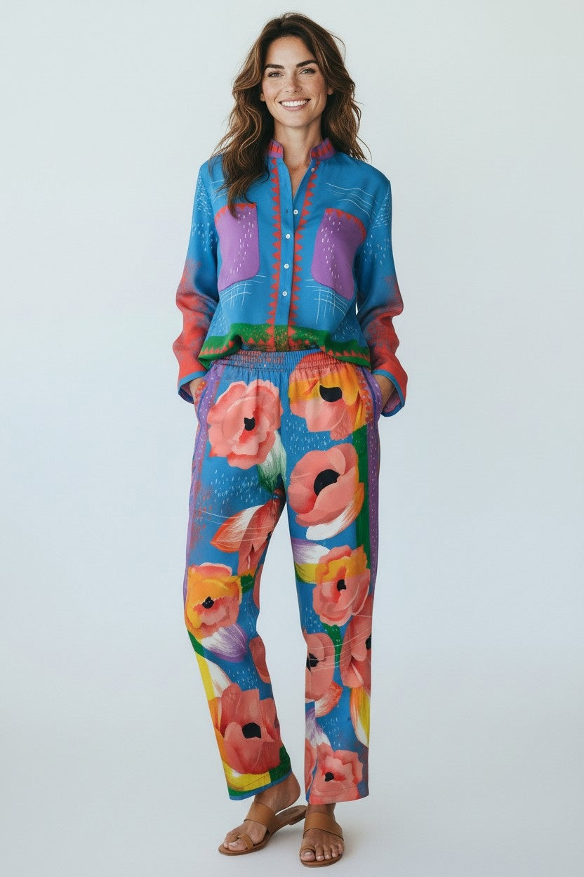 A person wears FRANCO FERRARI - SILK PRINT TROUSERS featuring a bold red, pink, and orange floral print on blue, paired with a matching top and brown sandals. Only the lower half of the body is visible.
