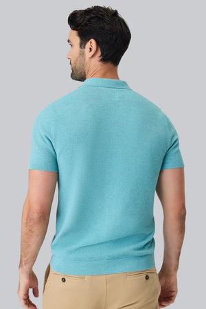 A man with short dark hair and a beard wears the MACK WELDON TECH LINEN SWEATER POLO in Deep Lagoon Heather, made with COOLMAX technology, and beige pants. He looks down, left arm bent, posed against a plain light gray background.