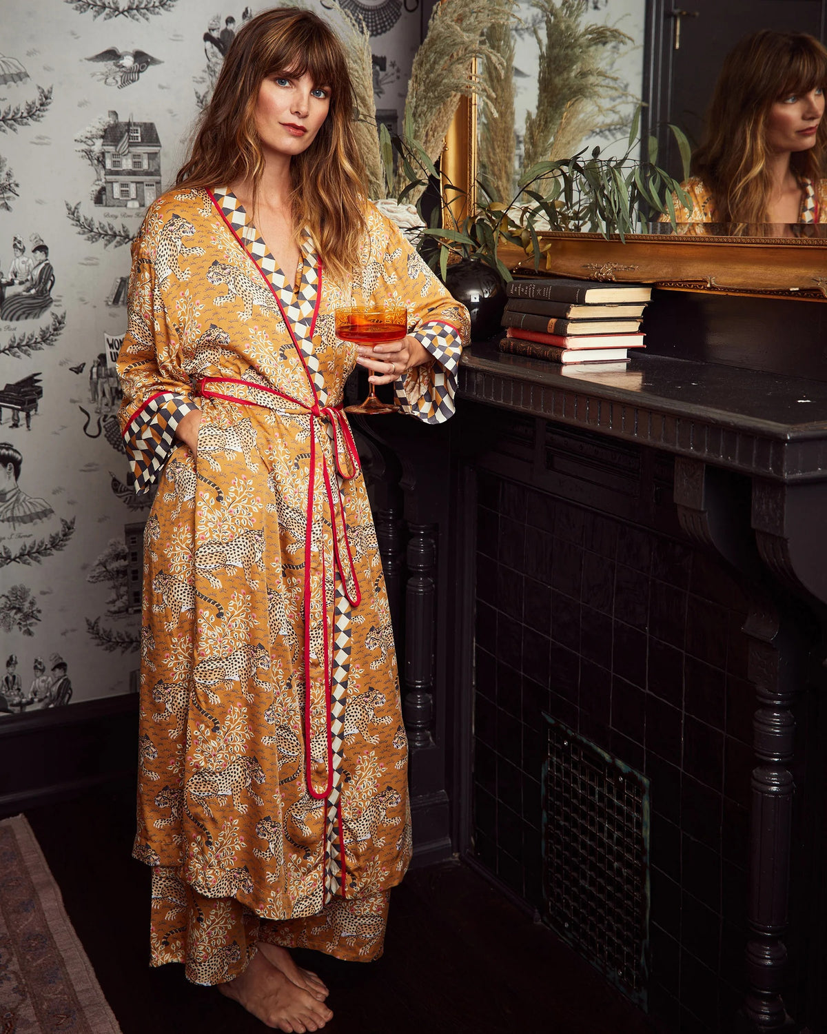 A woman stands barefoot, smiling, and wearing the PRINTFRESH Bagheera Eco Satin Robe in Tobacco—an eco-friendly viscose robe featuring a loose fit, bold patterns, and geometric trim. She is set against a white wooden panel wall with a beige carpet background.