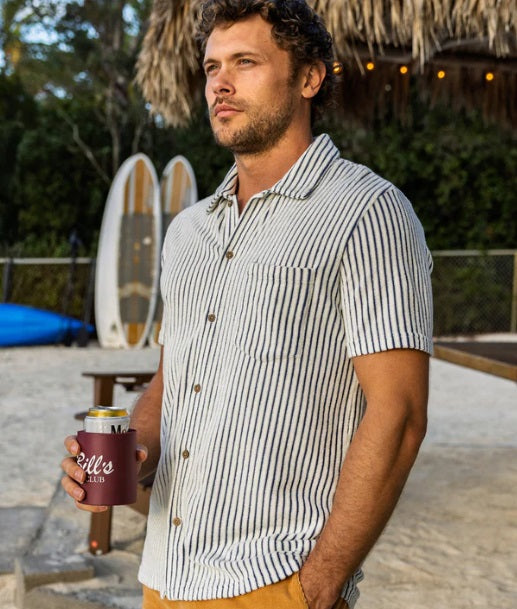 A man wears the CRIQUET Terrycloth Button Down Shirt in Striped Jacquard Natural—short-sleeved with vertical navy and white stripes and a chest pocket—paired with light pants for a breezy summer look.