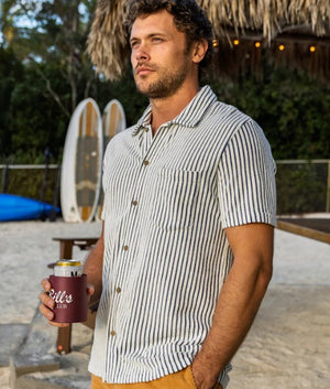 A man wears the CRIQUET Terrycloth Button Down Shirt in Striped Jacquard Natural—short-sleeved with vertical navy and white stripes and a chest pocket—paired with light pants for a breezy summer look.