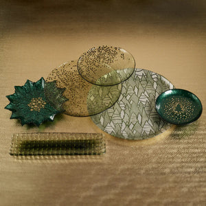 The DOTTED GLASS PLATE 8.25" by ZODAX is a round, shallow green plate with a textured surface and small, dark speckles on one side, perfect for serving and shown against a plain white background.