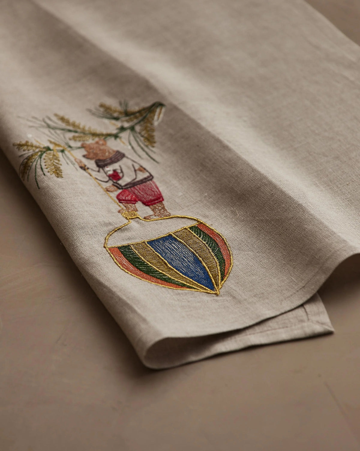 DECK THE HALLS BEAR LINEN TEA TOWEL featuring a bear in a sweater hanging a large, colorful, striped Christmas ornament from a pine branch on natural flax fiber.