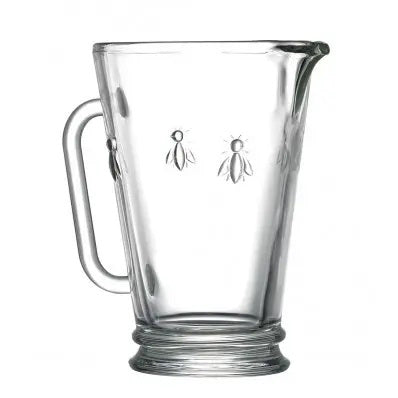 The BEE PITCHER by LA ROCHERE is displayed. This clear glass, all-purpose pitcher features an embossed bee design and is perfect for serving lemonade. It includes a handle on one side and a spout on the opposite side to ensure steady pouring. The bottom of the pitcher showcases a thicker glass base.