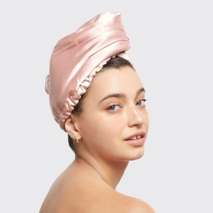 The image displays a beige box with the KITSCH Satin Wrapped Hair Towel inside. The packaging shows an illustration of the towel and a smiling person wearing it, emphasizing its benefits for reducing frizz in damaged, curly, or frizzy hair.