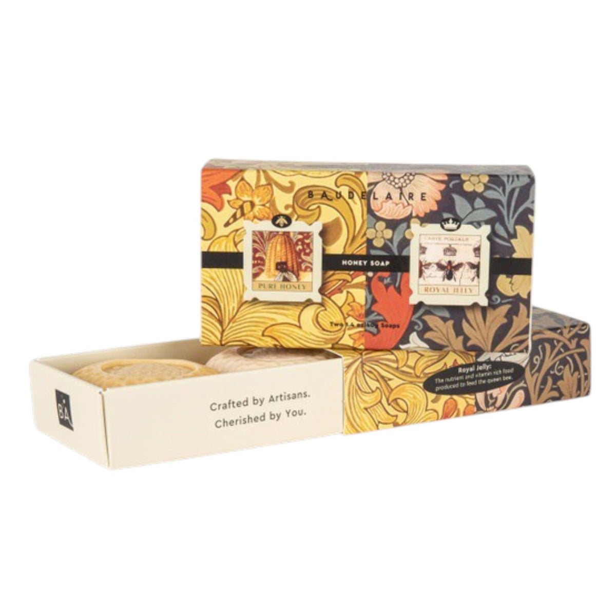Two BAUDELAIRE soap boxes with floral designs, one opened to reveal a honeycomb patterned soap bar inside. Labels show 'Pure Honey' and 'Royal Jelly'. Text on the side reads "Crafted by Artisans. Cherished by You." The BAUDELAIRE - PURE HONEY SOAP 1.4OZ MATCHBOX DUO is the perfect gift for any occasion. Background is plain white.