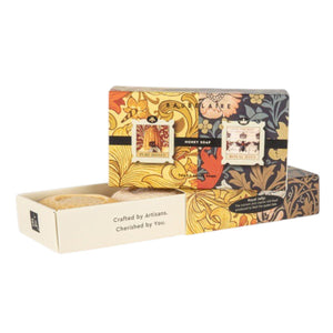 Two BAUDELAIRE soap boxes with floral designs, one opened to reveal a honeycomb patterned soap bar inside. Labels show 'Pure Honey' and 'Royal Jelly'. Text on the side reads "Crafted by Artisans. Cherished by You." The BAUDELAIRE - PURE HONEY SOAP 1.4OZ MATCHBOX DUO is the perfect gift for any occasion. Background is plain white.