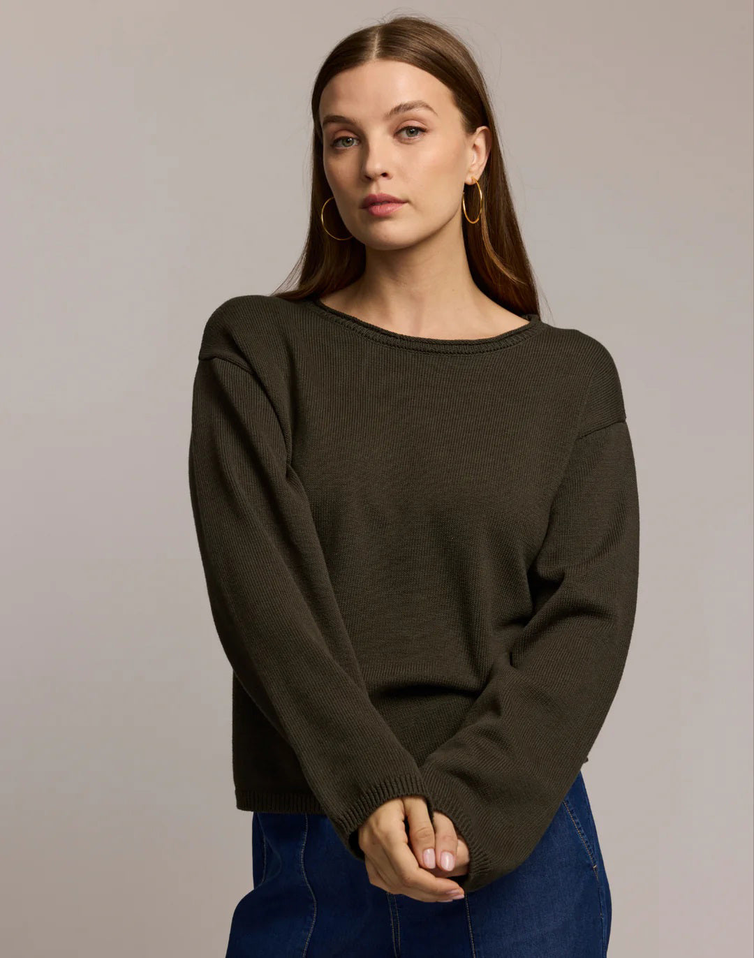 A woman with long brown hair and large hoop earrings models the HINSON WU AUDREY Long Sleeve Cotton Silk Sweater in tan with blue jeans, standing against a plain beige background, one hand raised near her shoulder.