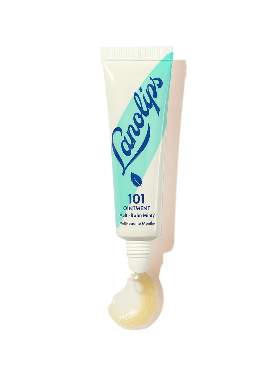 A woman holds a tube of LANOLIPS - 101 OINTMENT MULTI-BALM MINTY near her lips, dressed in a cream sweater against a blue background—highlighting LANOLIPS’ perfect minty hydration for dry lips.