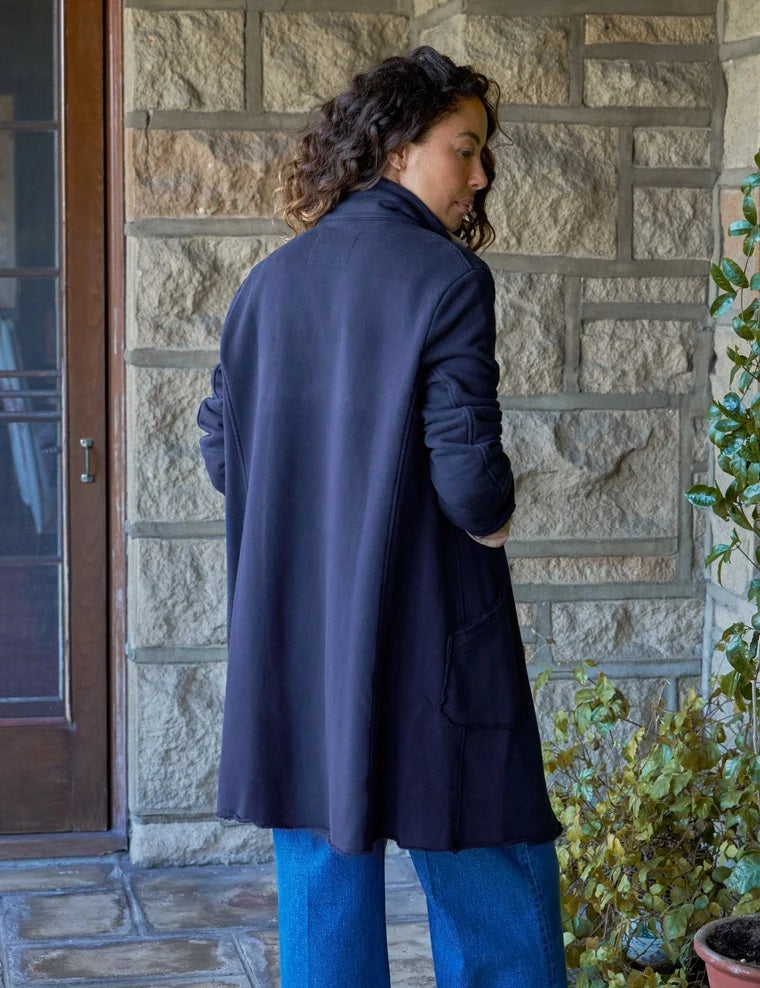 A woman stands indoors by a window, smiling with her hands in the pockets of a FRANK & EILEEN Belfast Sweatshirt Trench Coat in Classic Navy from the California Heritage Collection. She wears a tan shirt, wide-leg blue jeans, and brown boots. A wooden stool is positioned behind her.