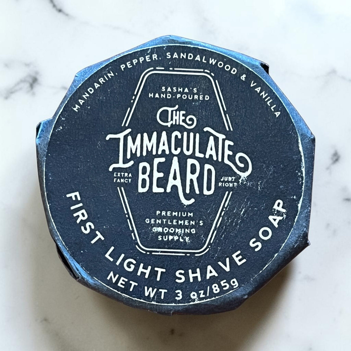 A round puck of THE IMMACULATE BEARD - SHAVE SOAP in First Light, ideal for sensitive skin, rests in a gray bowl. The black label features sandalwood, vanilla, and other notes from THE IMMACULATE BEARD.