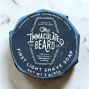 A round puck of THE IMMACULATE BEARD - SHAVE SOAP in First Light, ideal for sensitive skin, rests in a gray bowl. The black label features sandalwood, vanilla, and other notes from THE IMMACULATE BEARD.
