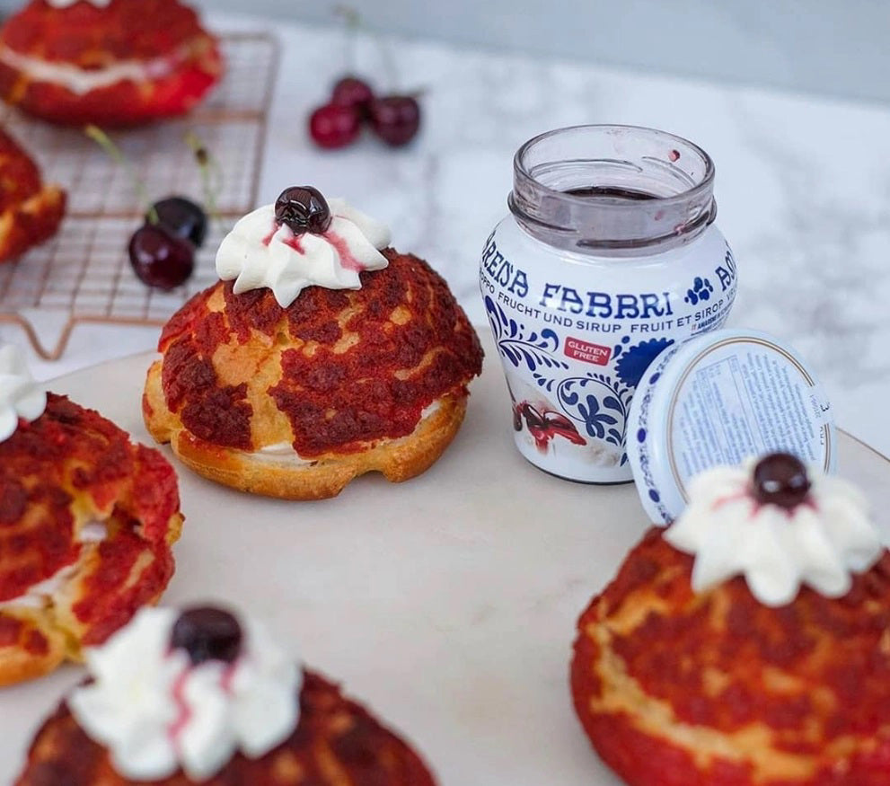 Cream puffs topped with whipped cream and a FABBRI - AMARENA CHERRIES IN SYRUP from GOURMET FOOD SOLUTIONS sit beside an open jar of the same cherries, while more cream puffs cool on a rack with fresh Italian tart cherries in the background.
