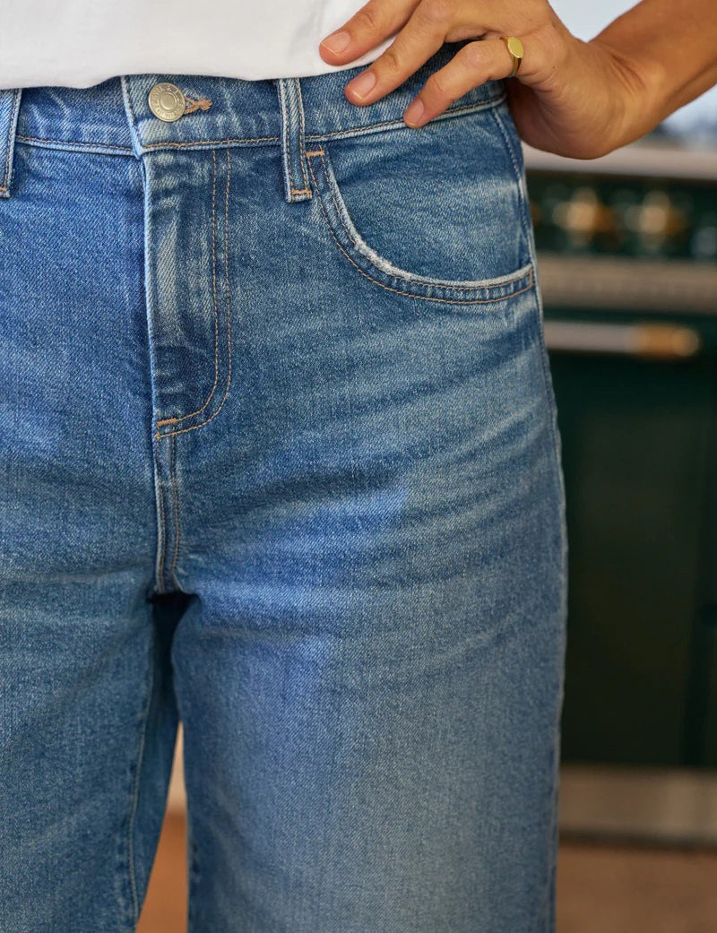 A person stands in a kitchen with their back to the camera, wearing FRANK &amp; EILEEN’s Brennan Everyday Barrel Jean in 1978 Vintage Wash, a white long-sleeve shirt, and brown shoes. The kitchen has light cabinets, a dark oven, and wood floors.
