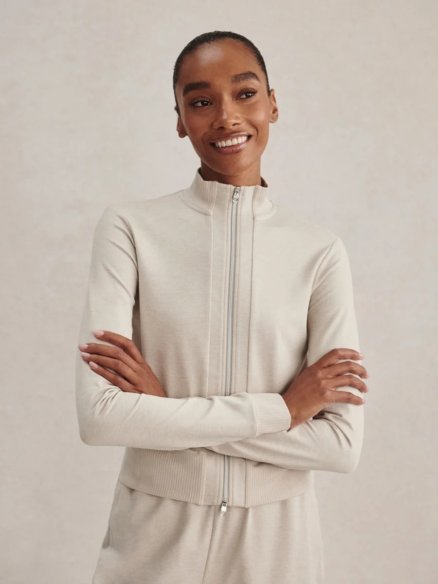 A person wears a light beige Varley Rufus Cropped Zip Through Sweat in Pebble Marl with a high collar and matching pants.