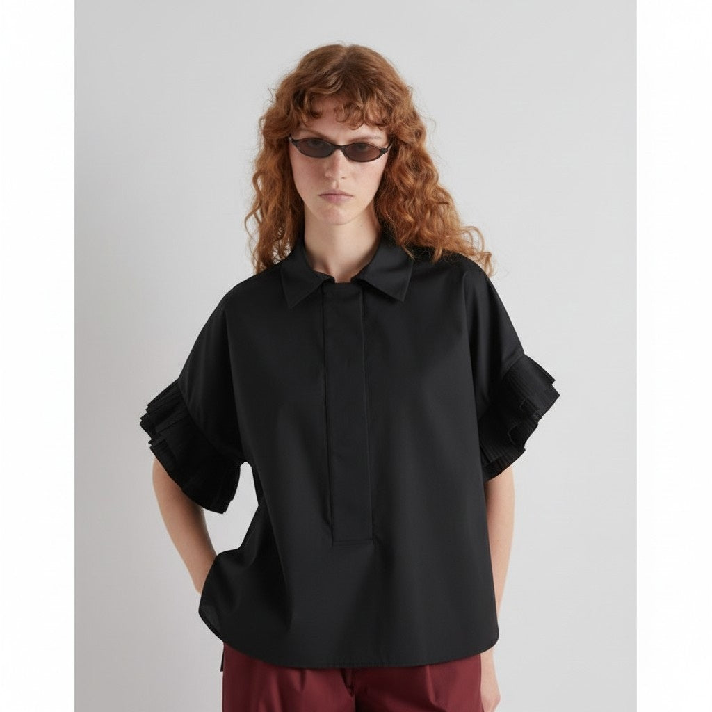 A woman with wavy red hair wears sunglasses and the MEIMEIJ - COTTON SHIRT IN BLACK by MEIMEIJ, featuring pleated ruffle sleeves and a boxy fit, paired with maroon pants against a plain light background.