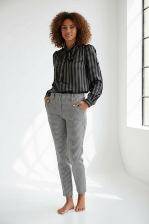 A woman with straight brown hair faces away in a HOPE FOR FLOWERS Tie Neck Blouse—black and white, vertically striped Tencel with puffed sleeves—paired with wide-leg plaid trousers. The plain gray background reflects sustainable style.