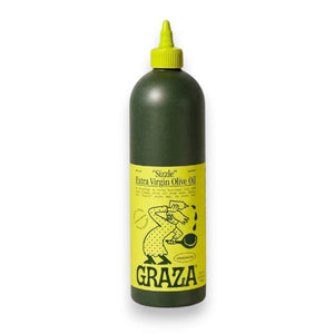A green bottle with a yellow label containing GRAZA - SIZZLE OLIVE OIL from the brand Graza is placed on a light-colored surface against a neutral background. The label, featuring an illustration of a person pouring olive oil and cooking-related text and graphics, proudly showcases its high-quality picual olive origins.