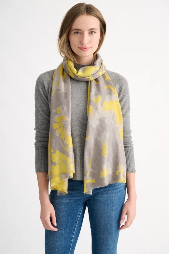 A woman wearing a gray sweater, blue jeans, and the KINROSS TAPESTRY SCARF IN CITRINE by KINROSS stands against a plain white background.