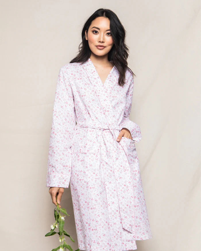 The PETITE PLUME - DORSET FLORAL ROBE by PETITE PLUME, displayed hanging, features a subtle, all-over pink floral pattern on a white background. This elegant robe is made from 100% cotton and includes long sleeves, a shawl collar, and a fabric belt tied at the waist—perfect for luxurious sleepwear.