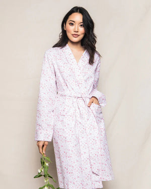 The PETITE PLUME - DORSET FLORAL ROBE by PETITE PLUME, displayed hanging, features a subtle, all-over pink floral pattern on a white background. This elegant robe is made from 100% cotton and includes long sleeves, a shawl collar, and a fabric belt tied at the waist—perfect for luxurious sleepwear.