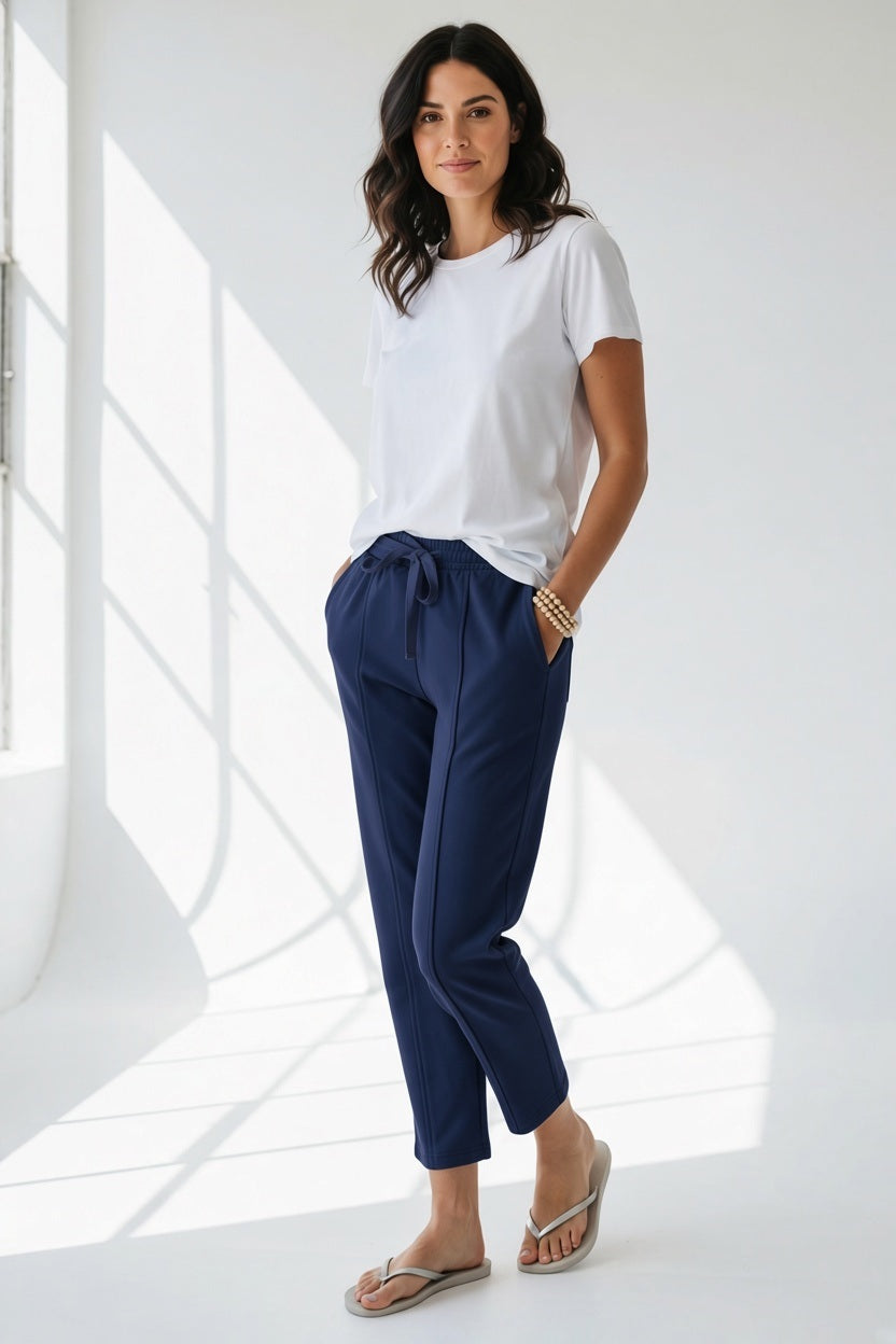 A person wears the MERSEA - FRENCHIE CIGARETTE PANT by MERSEA, paired with a white top and gray sneakers, standing on sandy beach ground with ocean waves and blue sky behind them, hands in their pockets.