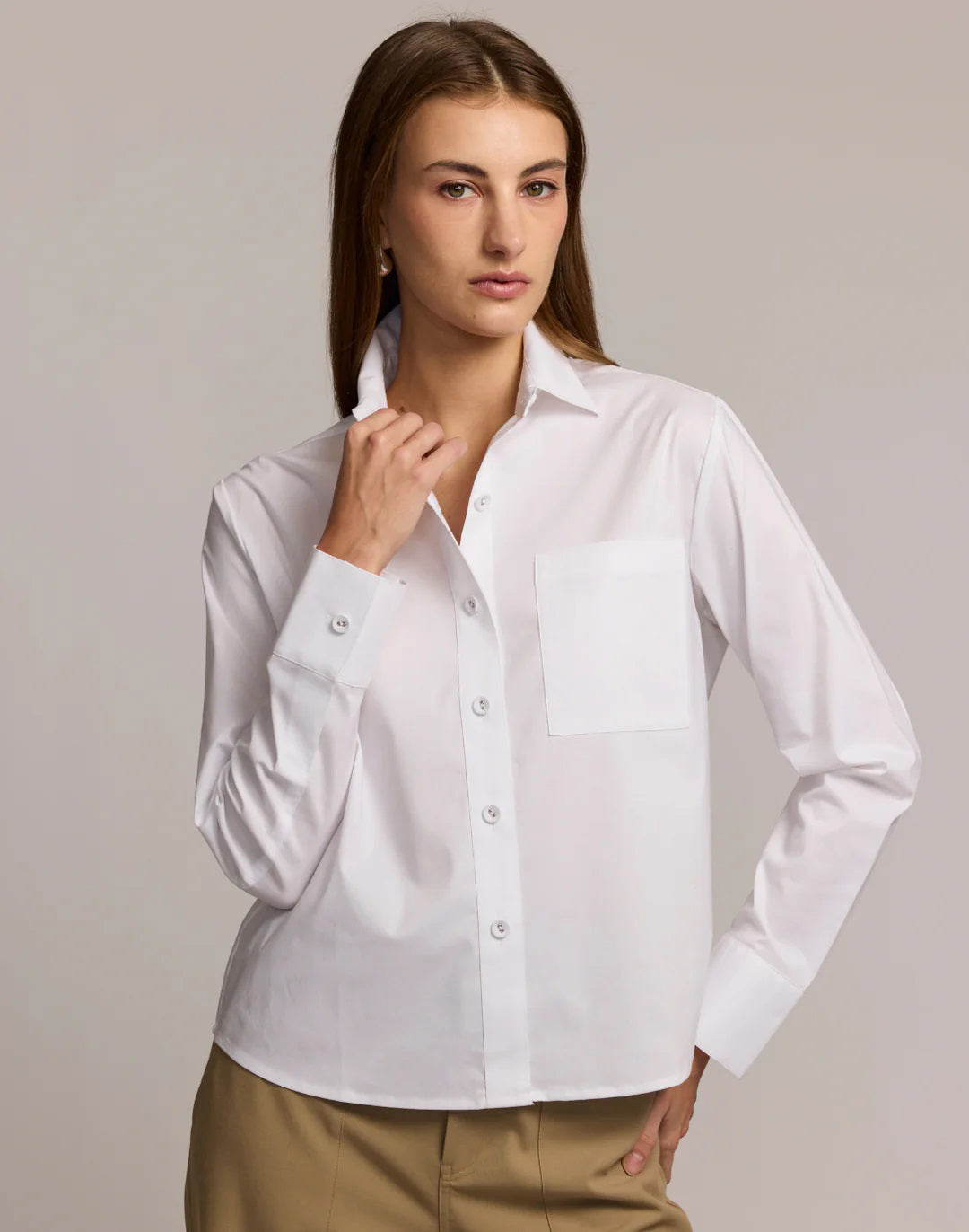 A woman wears the HINSON WU - VENUS Long Sleeve Untuckable Shirt in crisp white stretch cotton with a front pocket, paired with beige trousers, standing with hands in pockets against a neutral background.