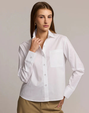 A woman wears the HINSON WU - VENUS Long Sleeve Untuckable Shirt in crisp white stretch cotton with a front pocket, paired with beige trousers, standing with hands in pockets against a neutral background.