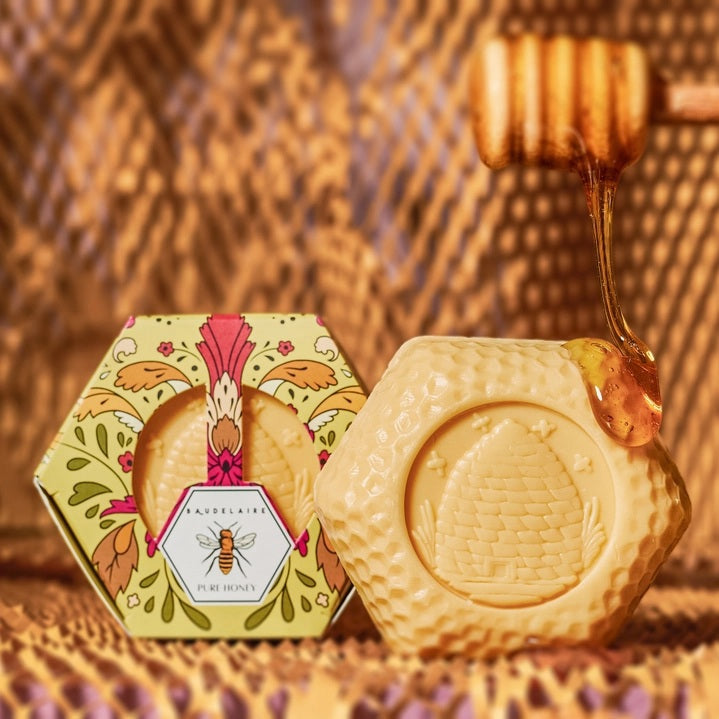 Hexagonal box of BAUDELAIRE - PURE HONEY SOAP 3.5 OZ, HEX BOX, featuring floral motifs and a bee illustration. The yellow and orange patterned packaging includes a top window that reveals the triple-milled BAUDELAIRE soap inside.