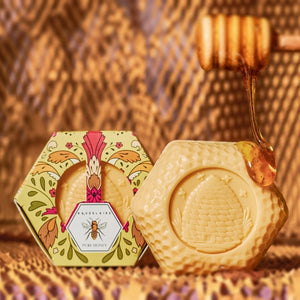 Hexagonal box of BAUDELAIRE - PURE HONEY SOAP 3.5 OZ, HEX BOX, featuring floral motifs and a bee illustration. The yellow and orange patterned packaging includes a top window that reveals the triple-milled BAUDELAIRE soap inside.