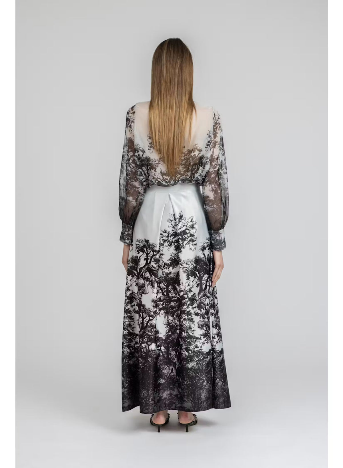 A woman wears a sheer, floral blouse paired with the MILVA MI - LONG MAXI SKIRT by MILVA—a shiny, high-waist skirt featuring a colorful floral print—and pointed black heels, posing against a simple gray studio backdrop.