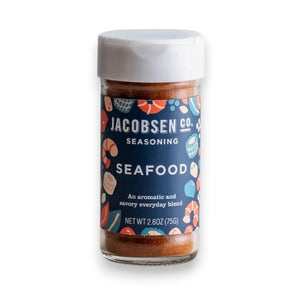 A clear spice jar with a white lid from JACOBSEN SALT CO., featuring a vibrant seafood-themed label for the JACOBSEN SALT - SEAFOOD SEASONING. The jar contains an orange-red blend made with sea salt, herbs, and spices—perfect for elevating your favorite seafood dishes.