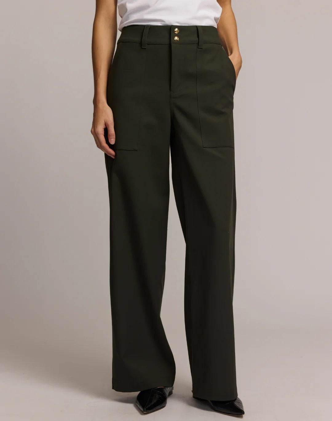 A person wears HINSON WU&#39;s KELLY TRAVELER&#39;S TWILL PANT in dark green with front pockets and gold buttons, styled with a white top and black pointed-toe shoes. Photo is cropped from shoulders down, highlighting this travel-ready look.
