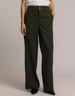 A person wears HINSON WU's KELLY TRAVELER'S TWILL PANT in dark green with front pockets and gold buttons, styled with a white top and black pointed-toe shoes. Photo is cropped from shoulders down, highlighting this travel-ready look.