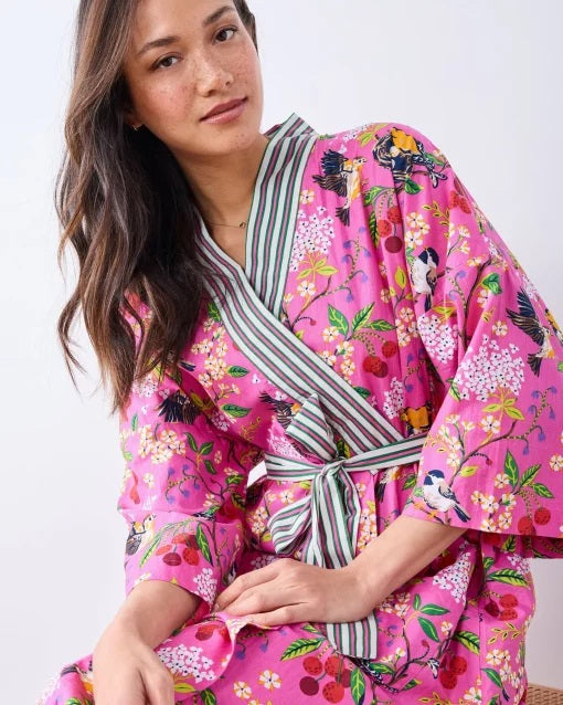 A person wearing a PRINTFRESH - BIRD & BLOSSOM LONG ROBE IN FUSCHIA, a long organic cotton robe with a bird and blossom pattern and green and white striped trim.
